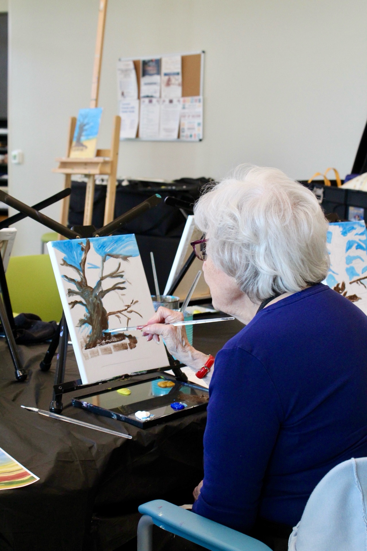 A senior painting at a table easel during a workshop session, with the demonstration painting on the easel behind her.